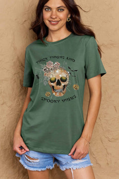 Green t-shirt with a skull surrounded by small Jackolanterns, batts, flowers and a spider. The text Reads Thick Thighs and Spooky Vibes