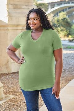 Woman wearing a green t-shirt and blue jeans standing outdoors with a bridge and trees in the background.