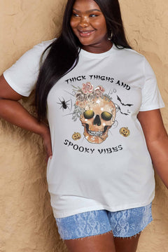 White t-shirt with a skull surrounded by small Jackolanterns, batts, flowers and a spider. The text Reads Thick Thighs and Spooky Vibes