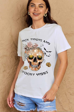 White t-shirt with a skull surrounded by small Jackolanterns, batts, flowers and a spider. The text Reads Thick Thighs and Spooky Vibes