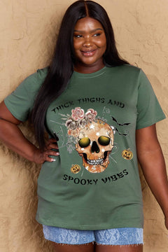 Green t-shirt with a skull surrounded by small Jackolanterns, batts, flowers and a spider. The text Reads Thick Thighs and Spooky Vibes