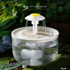 Cat water fountain with water flowing, surrounded by greenery clear bowl with white fountain head shaped like a daisy. Lettering says Little snail drinkingbowls