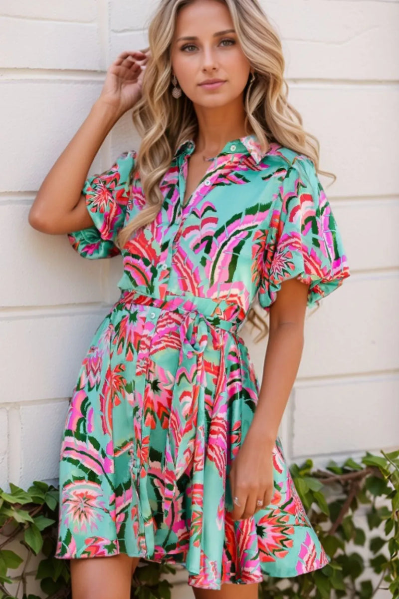 Abstract Print High Waist Buttoned Mini Dress. The background is a turquoise blue and the Abstract design gives n overall impression of pink, red, white and green flowers. Has a collar and small buttons down the front. full short skirt and full short sleeves, almost puffy. A fabric belt with same design is tied in bow offcenter of the waist.