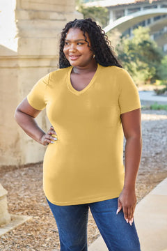 Woman wearing a yellow t-shirt and blue jeans standing outdoors with a bridge and trees in the background.