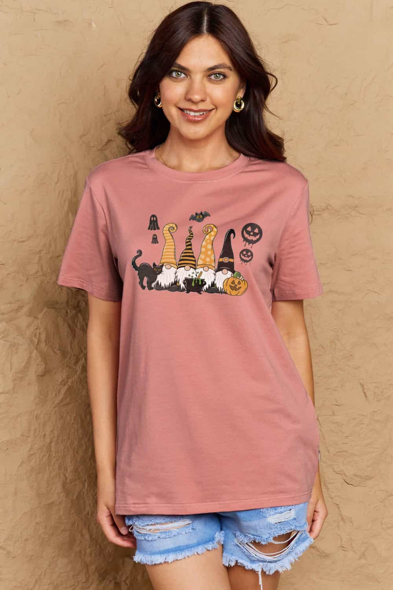  Pink t-shirt with Halloween-themed graphics. Four gnomes sitting behind a caldron with green brew bubbling out, a black cat with arched back on the right and a grinning jack o'lantern on the left with black 'sheet' ghosts, a bat and two black jack o' lantern silhouttes hovering above. Each gnome has a long pointy hat in either orange, black, or a combination of the two. The gnome on the left has a witch hat.