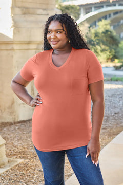 Woman wearing a coral t-shirt and blue jeans standing outdoors with a bridge and trees in the background.