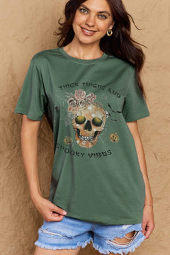 Green t-shirt with a skull surrounded by small Jackolanterns, batts, flowers and a spider. The text Reads Thick Thighs and Spooky Vibes