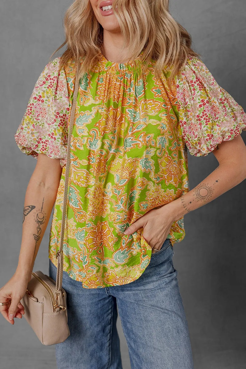 Multicolour Printed Contrast Bubble Sleeve Patchwork Blouse. The body is a bright green backgrouwn with orange and blue flowers, and the sleeves are a white background with smaller flowers in pink, red, yellow, with green foliage.
