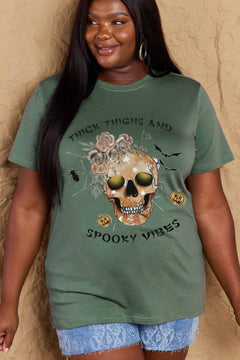 Green t-shirt with a skull surrounded by small Jackolanterns, batts, flowers and a spider. The text Reads Thick Thighs and Spooky Vibes