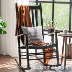 Black rocking chair with a plaid pillow and brown blanket in a room with a window and plants.