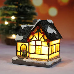 Decorative lit house with a snowy roof, glowing warmly against a blurred Christmas tree background.