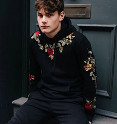 Person wearing a black hoodie with floral embroidery sitting against a dark door.