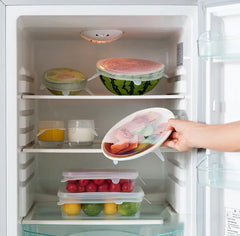 Refrigerator with food containers topped by reusable silicone stretch lids on shelves, hand holding a container.