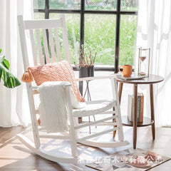 White rocking chair with a pink pillow and white blanket in a bright room with large windows.