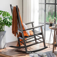 Black Rocking chair with an orange blanket in a bright room with plants and a table.