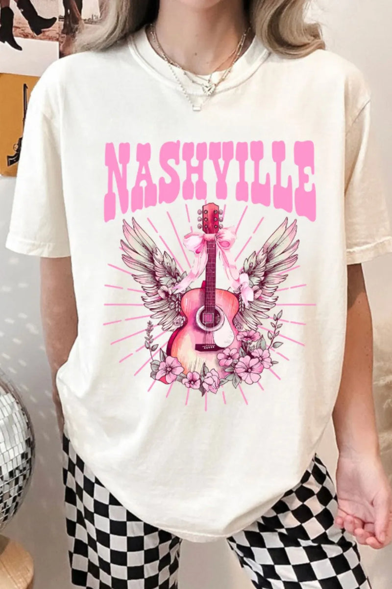 Creamy white Tshirt with short sleeves and round neck, the design is a Guitar standing on end, with the arm pointing up. There is a pink bow tied towards the top with long ribbon ends flowing down, pink and white wings extend out behind the guitar and the bottom is nestled in pink flowers that look similar to a carolina rose.Extending out all around the design are straight pink lines which imply rays of light. The caption above the design is in Pink Capital letters and says NASHVILLE