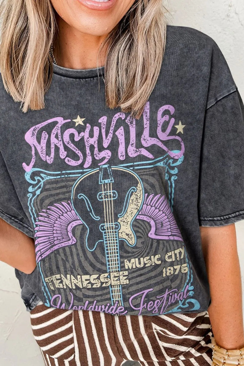 black Tshirt that is 'mineral washed' and faded to grey has Nashville in big Purple letters at top and World wide festival in the same purple at the bottom. Between those words is  a stylized blue box outline witha two tone guitar pointing down and purple wings spread out on each side, with the Words Tennessee Music City 1876 overlaid in tan.