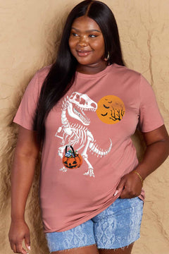 Pink short sleeved t-shirt with a dinosaur skeleton design on front. Dinosaur is holding a trick or treat jack o'lantern basket with candy in it and looking back at an orange moon with the silhouttes of bats and tree branches in front of it