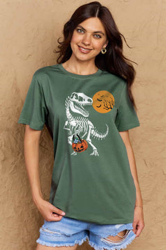 Green short sleeved t-shirt with a dinosaur skeleton design on front. Dinosaur is holding a trick or treat jack o'lantern basket with candy in it and looking back at an orange moon with the silhouttes of bats and tree branches in front of it