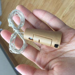 Hand holding a small cylindrical device with LED string attached on a wooden surface.