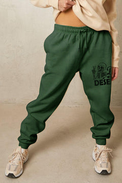 Green sweatpants with a graphic design on the left leg of the head and torso of a talking skeleton. Holding up it's right hand with the peace sign and the Slogan says "Have the day you deserve". Cuffed legs and elastic waist with drawstring and side pockets