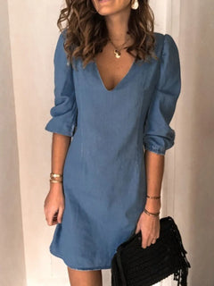 Woman wearing a blue dress holding a black clutch against a plain background
